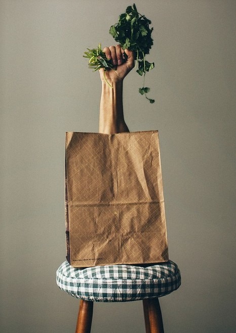 paper bag on a stool, hand coming out of the paper bag holding vegetables